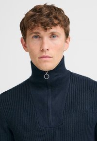 Navy ribbed sweater with a high neckline and half-zip closure. Features a textured knit pattern and a circular zipper pull.