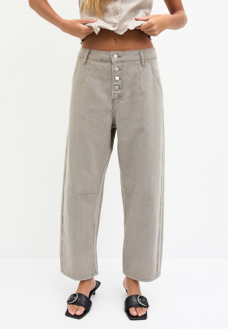 Femme portant un pantalon beige à fines rayures taille haute avec braguette à boutons et des sandales à talons noires à bout ouvert, ajustant une chemise courte beige.