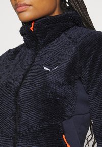 Navy blue fleece jacket with soft, textured surface and high collar. Features contrasting orange zipper accents and a logo detail.