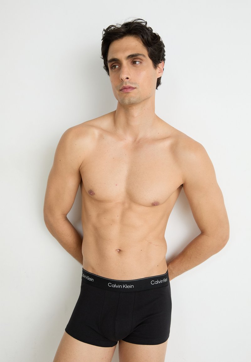 Zwarte katoenen boxershorts met een merk-elastische tailleband, gemodelleerd op een shirtloze mannelijke figuur tegen een lichte achtergrond.