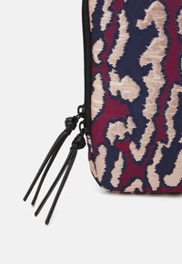 Close-up van een stoffen zak met een abstract patroon in rood, marineblauw en beige, en twee zwarte ritssluitingen met leren kwastjes met knopen.