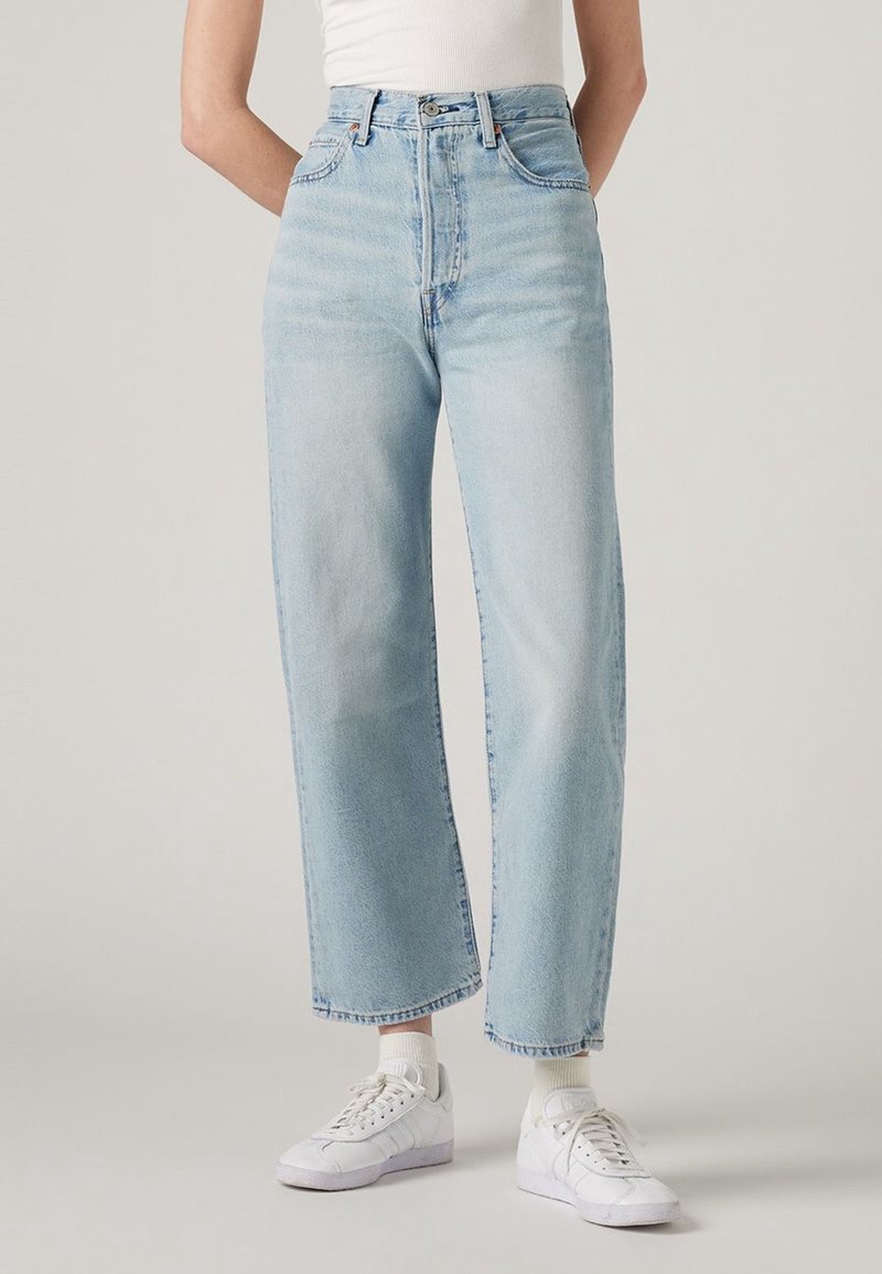 Personne portant un jean taille haute bleu clair à jambes larges, des chaussettes blanches, des baskets blanches et un haut blanc sans manches, debout les mains dans le dos.