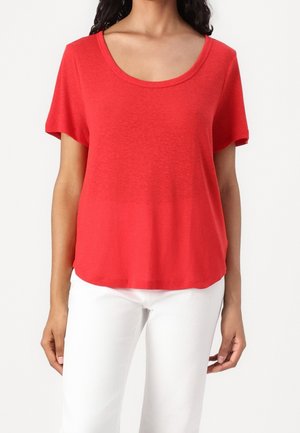 Woman wearing a bright red short-sleeve scoop neck top paired with white pants against a plain white background.