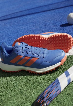 Blauwe en oranje sportschoenen op groen gras bij een blauw-witte veldhockeystick en een witte bal op een blauw speeloppervlak.