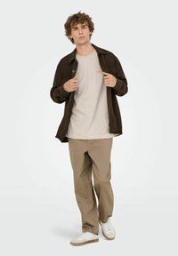 Chaqueta marrón de botones sobre una camiseta gris clara con un logo, pantalones beiges y zapatillas blancas con suela beige. Atuendo casual.
