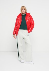 Doudoune rouge brillante avec un col haut, assortie à un haut zippé vert et un pantalon large crème avec une rayure verte. Baskets blanches.