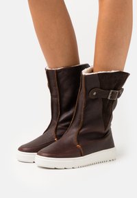 Brown leather ankle boots with a synthetic white sole, featuring a contrasting dark suede trim, decorative buckle, and warm lining.