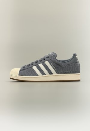 Baskets basses Adidas Superstar en gris foncé avec des rayures blanches, motif texturé, semelle crème et bout en caoutchouc.