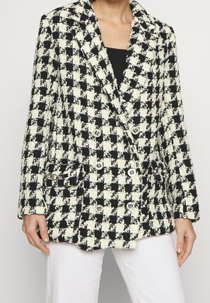 Frau trägt einen schwarz-weiß karierten Doppelreiher-Blazer mit silbernen Knöpfen und weiße Hose, steht vor einem schlichten Hintergrund.