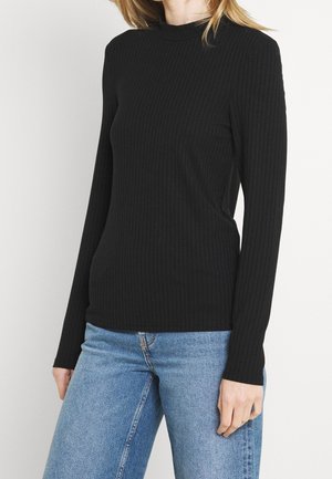 Schwarzes, geripptes Langarmoberteil mit hohem Kragen, tailliertem Schnitt und strukturiertem Stoff. Kombiniert mit hellblauen Jeans.