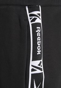 Svarta träningsbyxor med vit Reebok-logotape på sidan. Mjukt tyg, strömlinjeformad form och inga synliga detaljer eller dekorationer.