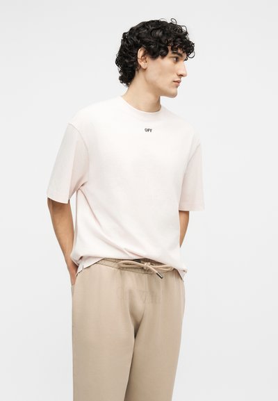Άνδρας με σγουρά μαλλιά που φοράει άσπρο φαρδύ T-shirt με το κείμενο "OFF" και μπεζ παντελόνι με κορδόνι, στέκεται με τα χέρια στις τσέπες.