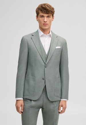 Junger Mann, der einen hellgrauen Anzug mit weißem Hemd und Einstecktuch trägt, steht vor einem schlichten hellen Hintergrund.