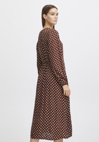 Robe à pois bruns avec manches longues, taille froncée, longueur midi, tissu lisse et col rond. Des pois blancs contrastent avec le fond.