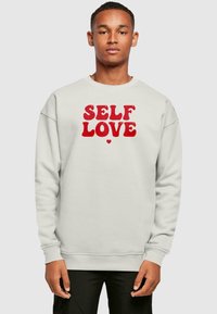 Felpa grigia chiara con la scritta "SELF LOVE" in bold rosso. Presenta un collo tondo, maniche lunghe e polsini e orlo a costine.