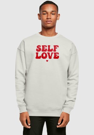 Lys grå trøje med "SELF LOVE" i fed rød skrift. Har rund hals, lange ærmer og ribbede manchetter og kant.