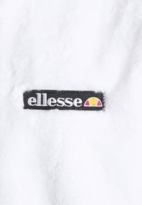 Patch logo Ellesse avec texte blanc et semi-cercle jaune-rouge sur tissu noir, cousu sur un matériau en polaire blanche douce.
