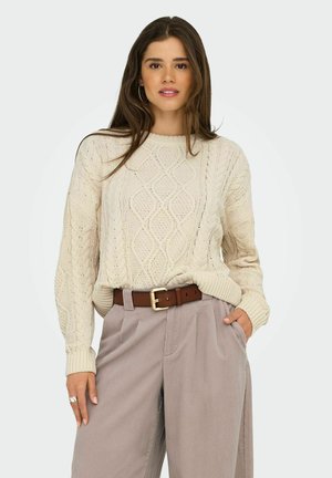 Maglione lavorato a maglia di colore crema con un motivo a diamante testurizzato, abbinato a pantaloni grigi chiari a pieghe e una cintura marrone con fibbia dorata.