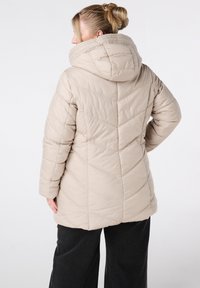 Beige pufferjack met capuchon, voorzien van chevronstiksels en een ontspannen pasvorm. Het materiaal lijkt zacht en textuur te hebben, geschikt voor koud weer.