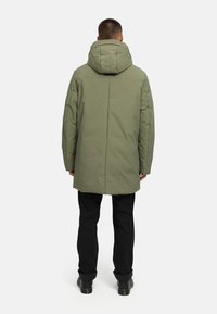 Olivovozelený zateplený kabát s kapucňou, s rovný strihom a detailami švov. Čierne nohavice a čižmy dopĺňajú outfit.