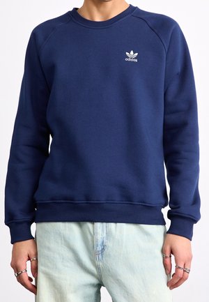Person trägt einen dunkelblauen Adidas-Pullover mit weißem Logo auf der Brust und hellblaue Jeans, zeigt den Oberkörper und die Hände mit Ringen an den Fingern.