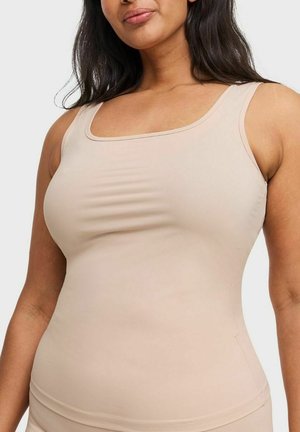 Vrouw draagt een mouwloos beige topje, afgebeeld van schouders tot heupen, tegen een effen lichte achtergrond.