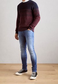 Hombre con un suéter de bloques de color, jeans ajustados de color azul claro y zapatillas altas negras, de pie sobre un suelo de madera con la mano en el bolsillo.