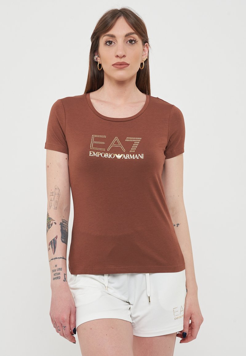 T-shirt marrone a maniche corte realizzata in tessuto morbido, con logo dorato "EA7 EMPORIO ARMANI" impreziosito da strass. Collo rotondo semplice.