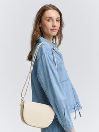 Borsa a tracolla beige con forma curva, texture liscia e tracolla regolabile, abbinata a una giacca in denim blu chiaro.