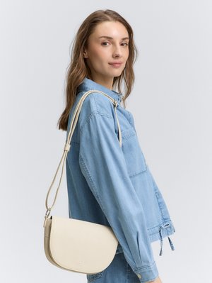 Beige schoudertas met een gebogen vorm, een gladde textuur en een verstelbare strap, gecombineerd met een lichtblauwe spijkerjack.
