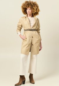 Beige trenchcoat met een riem, voorzien van twee voorzakken en een reverskraag. Gedragen over een lichte, textuur-geweven trui en witte broek met wijde pijpen.