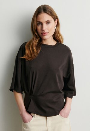 Donna con capelli castano chiaro ondulati, che indossa orecchini a cerchio d'argento, una maglietta nera corta e larga e pantaloni beige chiaro con le mani nelle tasche.