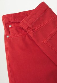 Jean en coton rouge avec une texture lisse, présentant des accessoires en métal argenté, une fermeture à bouton et des poches avant.