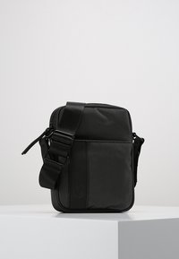 Black messenger bag with a textured fabric, adjustable strap, and a front pocket with a smooth leather accent. Compact rectangular shape.
