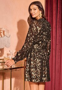 Černé šaty s metalickými leopardími skvrnami, dlouhými rukávy a nařaseným pasem; lehká průhledná tkanina; doplněné oversized náušnicemi.