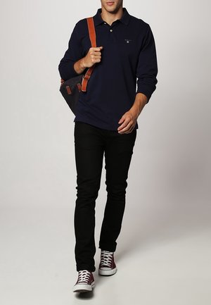 Polo shirt à manches longues bleu marine, pantalon slim noir et baskets bordeaux ; portant un sac noir avec une sangle marron ; cadre intérieur.