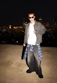 Siva široka jakna preko bele majice, uparena sa širokim sivim farmerkama i crnim platformama. Plava karirana košulja je vezana u struku.