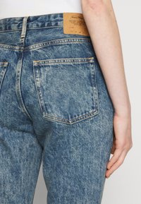 Zadní pohled na modré džínové kalhoty s vysokým pasem, dvěma zadními kapsami a koženou značkovou nášivkou na pase.