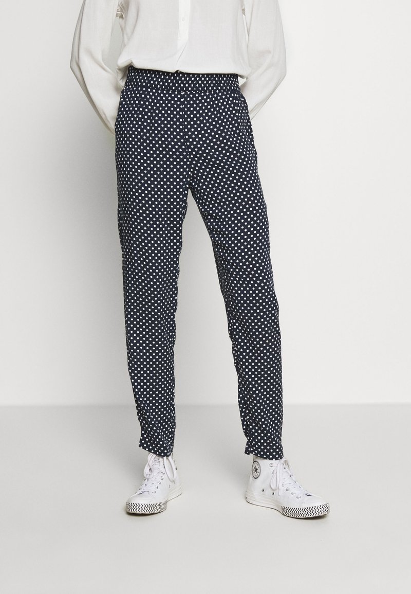 Personne portant un pantalon bleu marine à pois blancs, des baskets montantes blanches et une chemise blanche à manches longues rentrée, debout devant un fond uni.