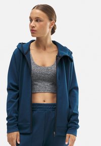 Chaqueta con capucha y cremallera en tejido azul oscuro, con un diseño deportivo que incluye una camiseta corta gris debajo y pantalones de cintura elástica.
