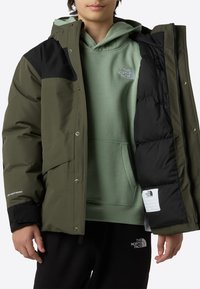 Olivengrønn isolert jakke med svarte detaljer, frontglidelås og trykknappslukking; kombinert med en lys grønn hettegenser og svarte bukser.