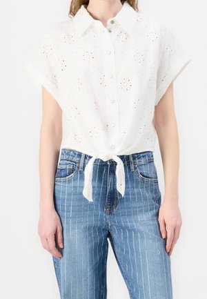 Biała koszula z krótkim rękawem zapinana na guziki, z ażurowym kwiatowym wzorem, związana w talii, w połączeniu z niebieskimi, wysokim stanem dżinsami z pionowymi białymi paskami.