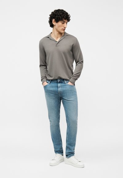 Νεαρός άνδρας που στέκεται με τα χέρια στις τσέπες, φοράει γκρι μακρυμάνικη πόλο, ανοιχτό μπλε τζιν και λευκά αθλητικά παπούτσια, κοιτάζοντας προς τα κάτω και προς τα αριστερά.