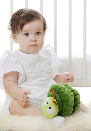 Toddler in white dress sitting on soft rug, holding a green plush caterpillar toy in bright, softly lit room.