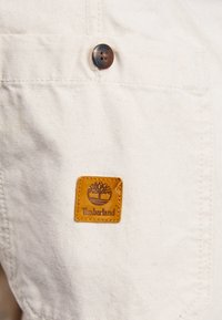 Hvidt stoflomme med en mørkebrun knap og et kvadratisk, syet lædermærke med et trælogo og "Timberland" tekst.