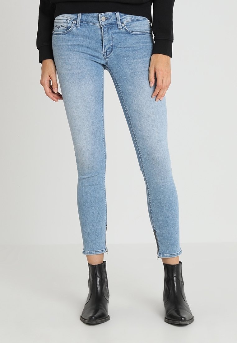 Jeans ajustados de color azul claro con cremalleras en los tobillos, usados con botas negras hasta el tobillo y una camiseta de manga larga negra, mostrados de la cintura a los pies.