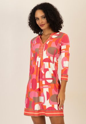 Woman wearing a colorful geometric patterned knee-length dress with three-quarter sleeves and embroidered neckline and cuffs.