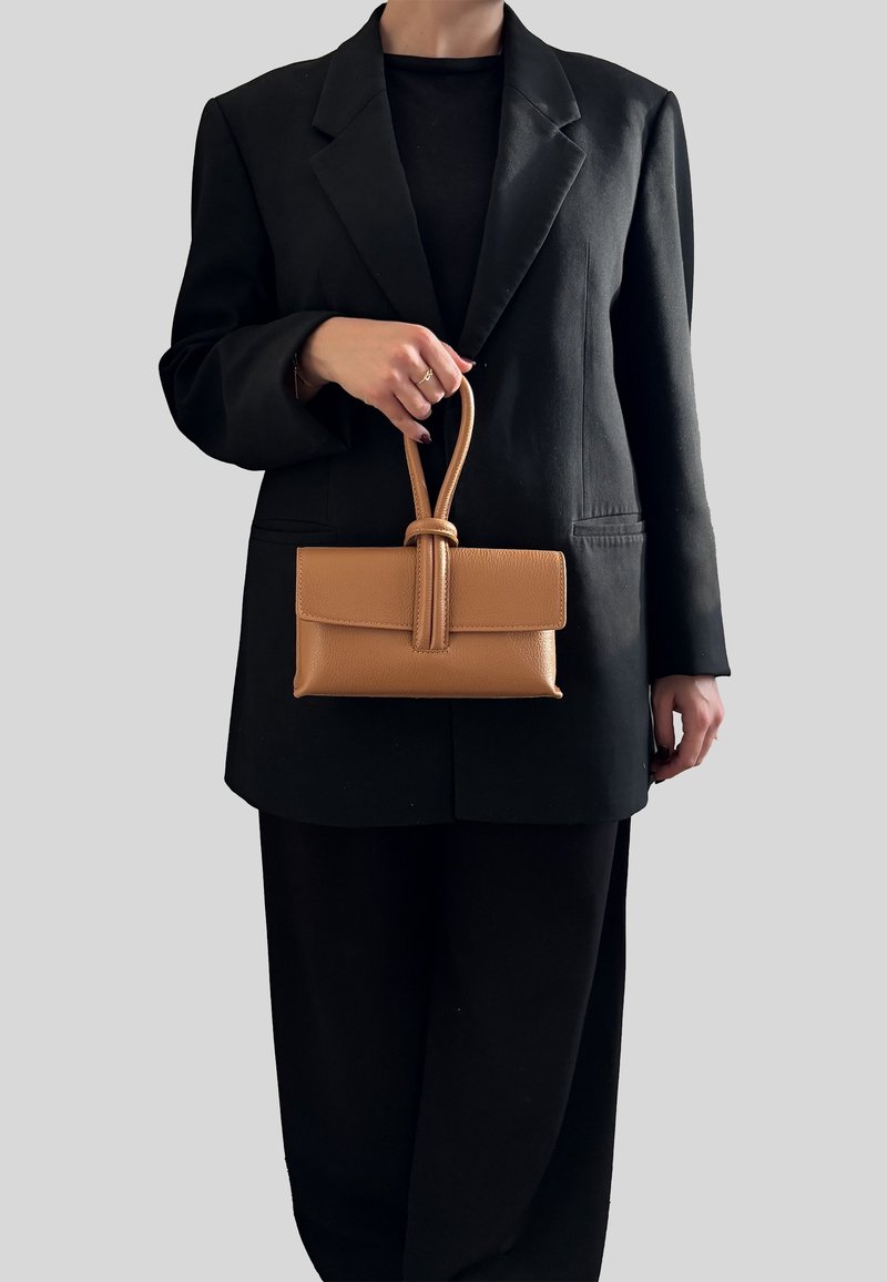 Femme portant un blazer noir et un pantalon, tenant un sac à main rectangulaire en cuir marron clair avec une poignée en boucle devant un fond gris.