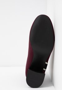 Semelle d'une chaussure bordeaux avec talon noir et taille 37 marquée, positionnée verticalement contre un fond blanc d'angle.