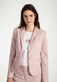 Lichtroze getailleerde blazer met twee knopen, revers en textuurstof. Draagt over een wit t-shirt met een bedrukt ontwerp.